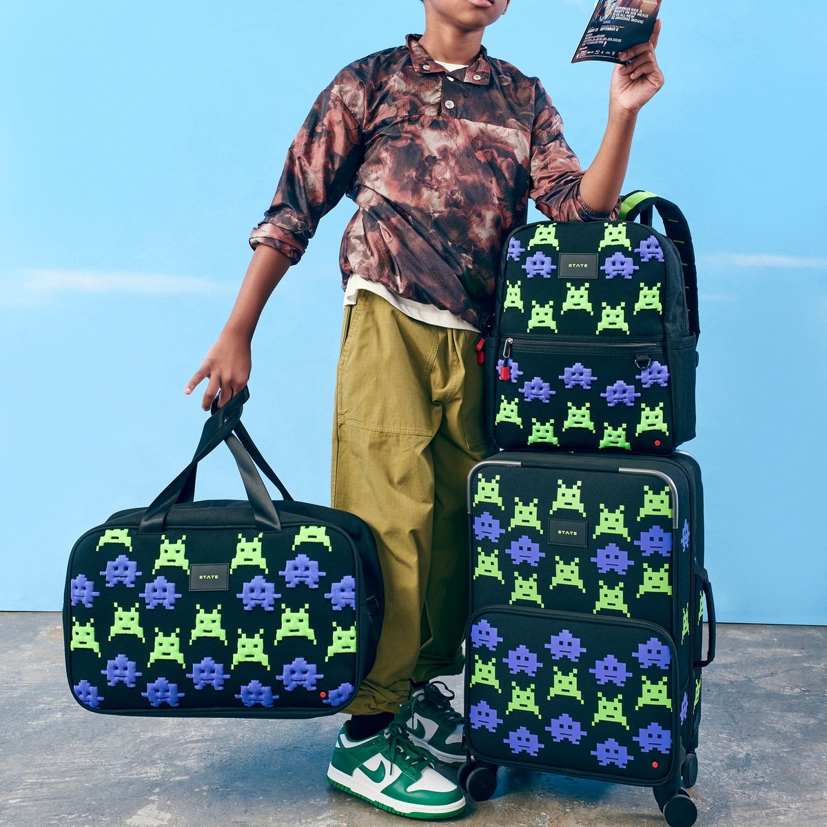 Boy holding STATE Bags Rockaway duffle and leaning on kane bacpack and logan carry-on arcade